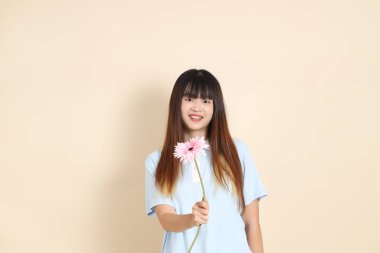 Açık mavi polo tişörtlü, beyaz şortlu, bej arka planda çiçek tutan orta boy bir Asyalı kadın resmi. Üniversite öğrencisi, Gen Z