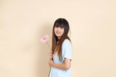 Açık mavi polo tişörtlü, beyaz şortlu, bej arka planda çiçek tutan orta boy bir Asyalı kadın resmi. Üniversite öğrencisi, Gen Z