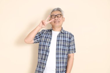 Gözlüklü, barış işareti, V işareti, iki parmak, günlük ekose gömlek, şort ve spor ayakkabı giyen, bej arka planda izole Asyalı bir son sınıf öğrencisi portresi. Yaşlı erkek yaşam tarzı konsepti.
