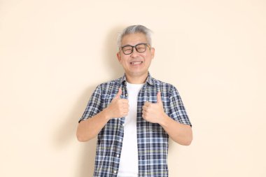 Gözlüklü, üst düzey Asyalı bir adamın portresi, tamam ya da onaylandı, bej arka planda izole edilmiş günlük ekose gömlek, şort ve spor ayakkabı giyiyor. Yaşlı erkek yaşam tarzı konsepti.