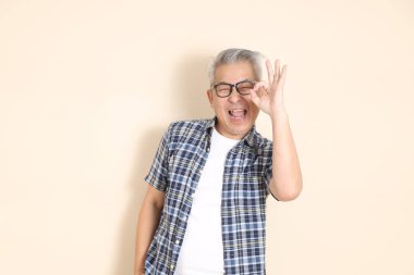 Gözlüklü, üst düzey Asyalı bir adamın portresi, tamam ya da onaylandı, bej arka planda izole edilmiş günlük ekose gömlek, şort ve spor ayakkabı giyiyor. Yaşlı erkek yaşam tarzı konsepti.