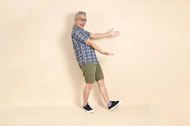 Gözlüklü bir Asyalı 'nın portresi, bej arka planda izole edilmiş, günlük ekose gömlek, şort ve spor ayakkabı giyiyor. Yaşlı erkek yaşam tarzı konsepti.