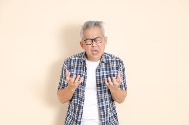 Gözlüklü, hayal kırıklığına uğramış, ağlayan, günlük ekose gömlek, şort ve spor ayakkabı giyen, bej arka planda izole Asyalı bir adamın portresi. Yaşlı erkek yaşam tarzı konsepti.