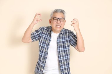 Gözlüklü, sinirli ve çığlık atan, günlük ekose gömlek, şort ve spor ayakkabı giyen, bej arka planda izole edilmiş Asyalı bir son sınıf öğrencisinin portresi. Yaşlı erkek yaşam tarzı konsepti.