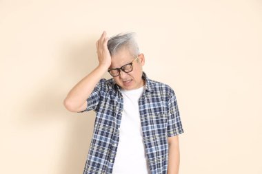 Gözlüklü, baş ağrısı ya da stresi olan, ekose gömlek, şort ve spor ayakkabı giyen, bej arka planda izole Asyalı son sınıf öğrencisinin portresi. Yaşlı erkek yaşam tarzı konsepti.