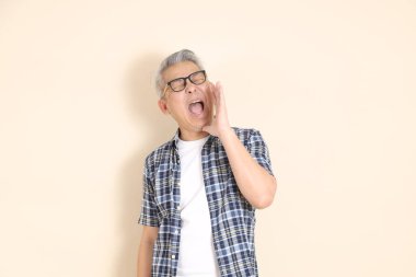 Gözlüklü, bağıran, megafon tutan, günlük ekose gömlek, şort ve spor ayakkabı giyen, bej arka planda izole Asyalı son sınıf öğrencisinin portresi. Yaşlı erkek yaşam tarzı konsepti.