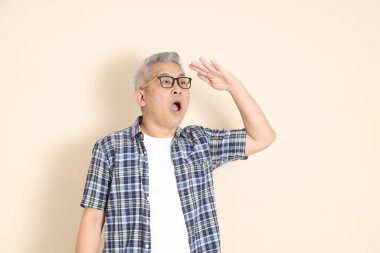 Gözlüklü Asyalı son sınıf bir adamın portresi, uzaklara bakıyor, ekose gömlek, şort ve spor ayakkabı giyiyor, bej arka planda izole edilmiş. Yaşlı erkek yaşam tarzı konsepti.