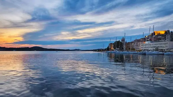 Gün batımında deniz manzarası. Güneş bir körfezin sakin sularında batıyor. Bağlı tekneler ve binalar denize yansıyor..