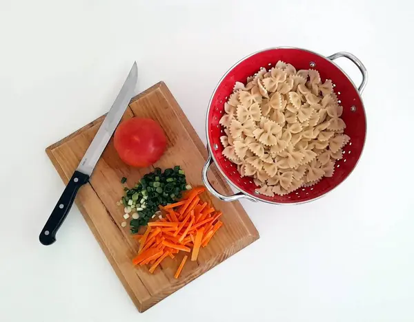 Kırmızı bir süzgeçte pişirilmiş farfalle makarna ve bıçak ve sebzeli bir kesim tahtası (taze soğan, havuç, domates).