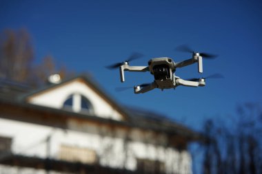 Tek kişilik bir evin önünde kameralı bir dron geziniyor. Dengeleyici 4K ile etiketlenmiş. 250 gramın altında hafif bir dron. Beyaz bir aile evinin ve mavi gökyüzünün bulanık arkaplanı..