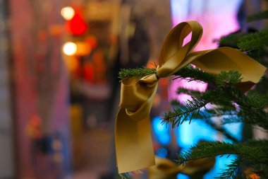 Bir Tannenbaum dalında süslenmiş altın kumaştan bir kurdeleyle tatil ve Noel alışverişini görselleştirin. Arkaplanda kırmızı, pembe, mavi ve sarı renkli canlı renkler var. Büyüleyici bir bokeh etkisi yaratıyor.