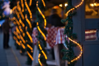 Bu dikey formatta kendinizi büyüleyici Noel ışıklarının ışıltısına daldırın. Fotoğrafta, suni çam çelenkleriyle süslenmiş bir panç standının gri ahşap cephesinde dolanan bir ışık tüpü görülüyor. Kompozisyon foku