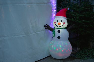 Kırmızı bir Noel şapkası takmış şişme bir kardan adamın kaprislerine kucak aç, içten içe parlak LED ışıklarıyla aydınlatılıyor. Dost canlısı, güler yüzlü ve kollarını açmış bu neşeli figür sağda koyu bir çam ağacının yanında duruyor. 