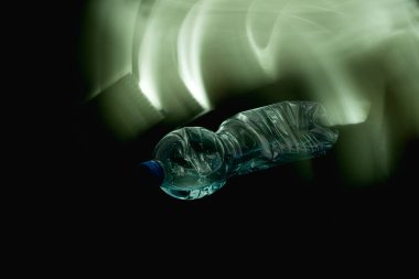 Kırık plastik bir şişe kısmen suyla dolu, siyah arka planda yatay olarak yatıyor, 45 derecelik açıyla vida kapağı kameraya bakıyor. Plastik şişe, plastik bir süngerle hafif boya tekniği kullanılarak aydınlatılıyor.