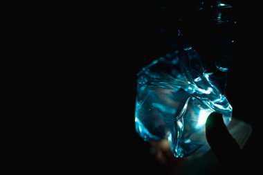 Şeffaf bir plastik şişe sıkıştırılmış ve su içeriyor, zar zor görünen bir elle tutuluyor. Koyu siyah bir arka planda, ışıkla aydınlatılmış. Dernekler arasında plastik atık, su kirliliği, su kıtlığı, sudaki plastik ve çevresel kirlilik yer alıyor