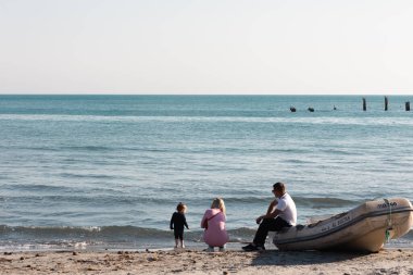 Durres, Arnavutluk - 13 Nisan 2024 - Durres gezinti güvertesinde küçük bir teknenin yanında duran bir ailenin fotoğrafı.