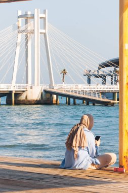 Arnavutluk 'un Durres kentinde 13 Nisan 2024' te deniz kenarındaki ahşap bir platformda telefonuyla oturan örtülü bir kadının fotoğrafı. Yüksek kalite fotoğraf