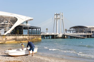 Durres, Arnavutluk - 13 Nisan 2024 - Durres gezinti güvertesinde kıyıda küçük bir tekne ve teknede küçük bir çocukla duran bir adamın fotoğrafı.