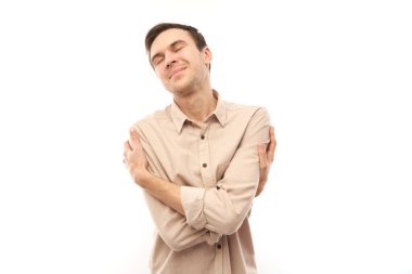 Beyaz stüdyo arka planında kendini kucaklayan ve gülümseyen genç esmer erkek. Kendini sevme ve kendini koruma kavramı