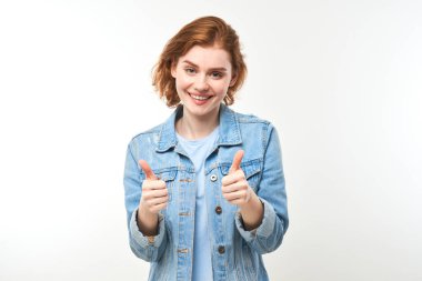 Genç kızıl saçlı kadın elini kaldırıyor ve beyaz arka planda izole kameraya gülümsüyor.