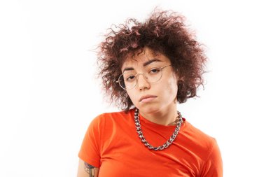 Beyaz stüdyo arka planında izole edilmiş, kıvırcık saçlı, dövmeli ve ciddi yüzlü havalı model kızın yakın plan portresi.