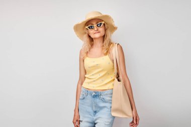 Sarı yaz kıyafetleri içinde mutlu sarışın olgun bir kadının portresi, hasır şapka, güneş gözlüğü ve alışveriş çantası beyaz stüdyo arka planında izole edilmiş bir şekilde gülümsüyor. Tatil ve seyahat kavramı