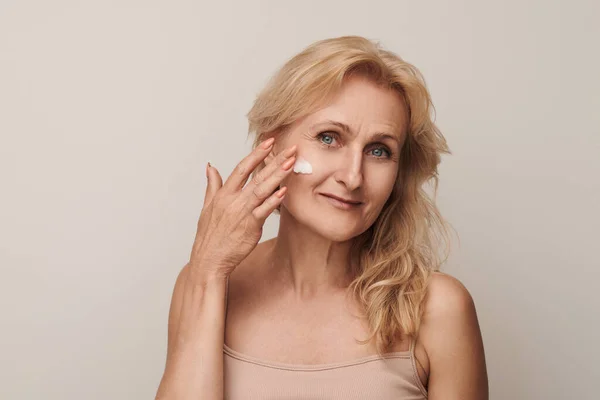 Portrait of luxurious looking 50s middle-aged mature blond woman smiling applying cream on face isolated on white studio background. Anti-aging skin care concept