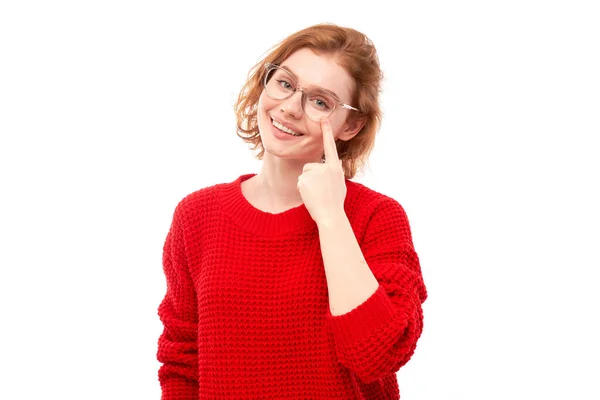 stock image Portrait of young woman in red sweater pointing finger at glasses on face isolated on white studio background. Vision check and loss of sight, search concept, eyewear shop