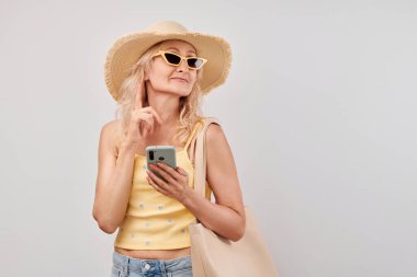 Saman şapkalı, sarı bluzlu, güneş gözlüklü, akıllı telefon ve alışveriş çantalı, beyaz stüdyo arka planında izole edilmiş, olgun sarışın kadın portresi.