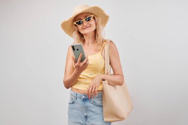 Saman şapkalı, sarı bluzlu, güneş gözlüklü, akıllı telefon ve alışveriş çantalı, beyaz stüdyo arka planında izole edilmiş, olgun sarışın kadın portresi.