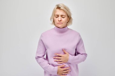 Portrait of young blonde woman suffering from stomach pain isolated on white studio background. Intestinal and stomach problems concep clipart