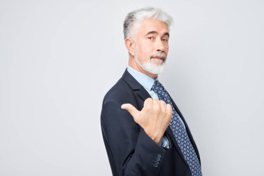 Gri saçlı, yetişkin bir adam 60 yaşında, iş elbisesi içinde, beyaz stüdyo arka grubunda izole edilmiş ürün ya da metin için boş alan göstererek parmağını bir kenara koyuyor.