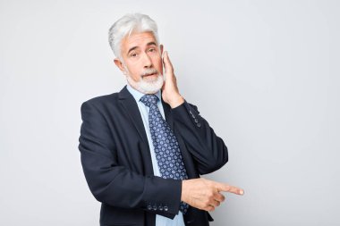 Gri saçlı, yetişkin bir adam 60 yaşında, iş elbisesi içinde, beyaz stüdyo arka grubunda izole edilmiş ürün ya da metin için boş alan göstererek parmağını bir kenara koyuyor.