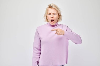 Genç, sarışın bir kadın, gösterişsiz bir şekilde işaret ediyor. Beyaz stüdyo arka grubunda boş bir alan ya da mesaj gösteriyor.