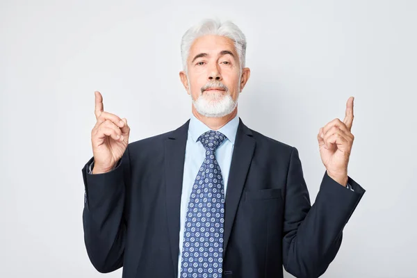 Gray-haired mature man 60 years old in business suit pointing up finger, demonstrating empty space for product or text isolated on white studio backgroun