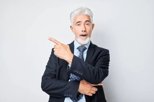 Gray-haired mature man 60 years old in business suit pointing aside finger, demonstrating empty space for product or text isolated on white studio backgroun