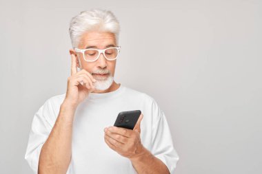 Beyaz tişörtlü gri saçlı adamın portresi cep telefonunda görünüyor ve düşünüyor. Akıllı telefonu olan kişi beyaz grupta izole edildi
