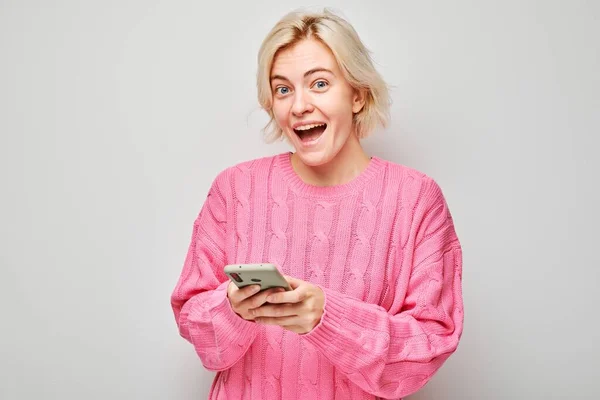 Portrait of young blond woman in pink sweater holding mobile phone in hand with happy smiling face. Person with smartphone isolated on white backgroun