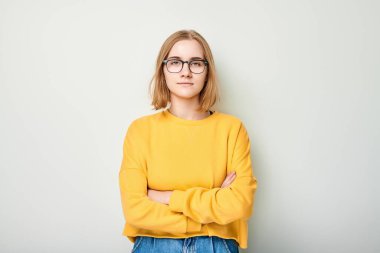 Gözlüklü ve sarı kazaklı, kolları arka planda çapraz duran kendinden emin genç bir kadın.