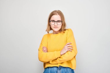 Gözlüklü ve sarı süveterli genç bir kadın kollarını gri bir arkaplanda kavuşturmuş duruyor..