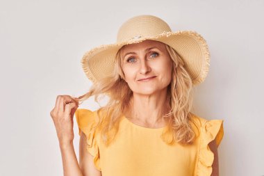 Sarı bluzlu ve hasır şapkalı gülümseyen kadın açık arkaplanda şakacı bir poz veriyor..