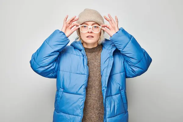 Young woman in blue puffer jacket and beanie adjusting glasses against a gray background.