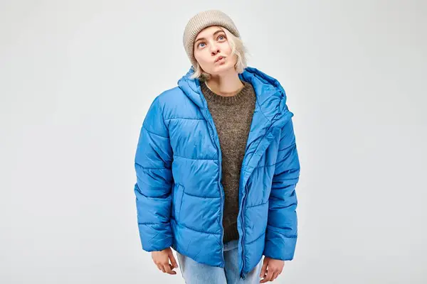 Young woman in blue puffer jacket and beanie posing with a curious expression on a plain background.