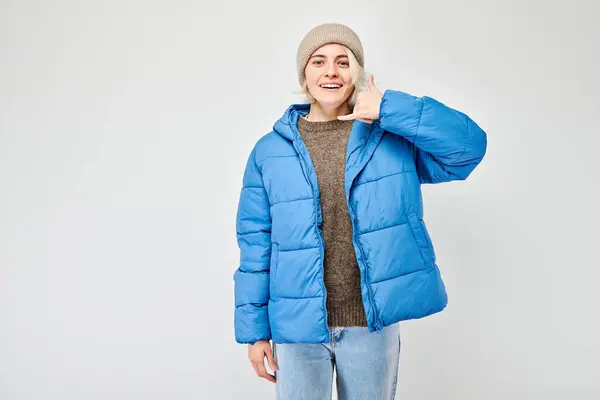 Smiling person in a blue puffer jacket and beanie making a call me gesture against a gray background