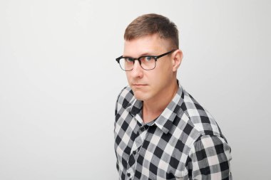 Gözlüklü ve kareli gömlekli şüpheci genç adamın portresi beyaz arka plandaki kameraya bakıyor.