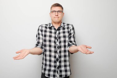 Karmaşık gömlekli genç adam beyaz arka planda omuzlarını silkiyor ve belirsizlik gösteriyor.