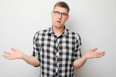 Karmaşık gömlekli genç adam beyaz arka planda omuzlarını silkiyor ve belirsizlik gösteriyor.