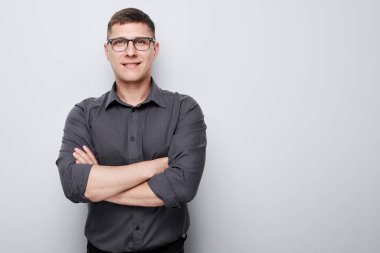 Gözlüklü, gri gömlekli, kolları çaprazlanmış, sade bir arka planda duran kendinden emin genç adam..