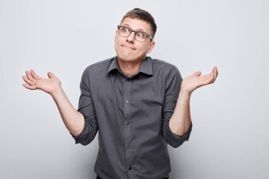 Kafası karışmış genç bir adam gri bir arka plana karşı omuz silkiyor.