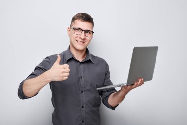 Gözlüklü gülümseyen adam dizüstü bilgisayarını tutuyor ve gri bir arka plana başparmak işareti yapıyor..